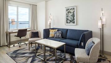 Ett modernt hotellrum med ett stort fönster, en blå soffa, ett litet bord, en stol och en golvlampa.