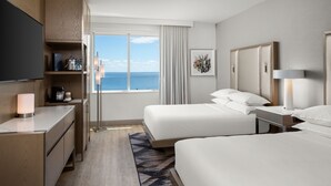 Et hotelværelse med to senge, et fjernsyn, et skrivebord med lampe, kaffemaskine og havudsigt.