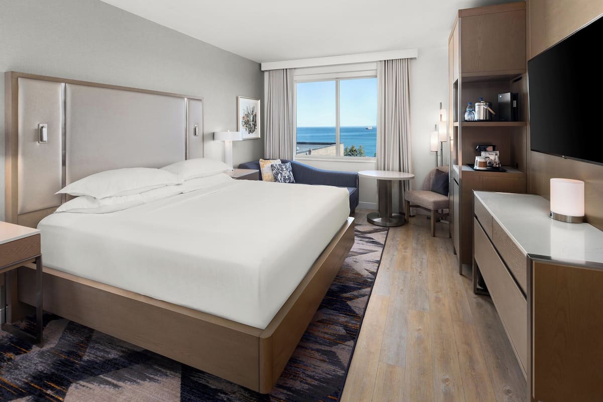 A modern hotel room with a large bed, a desk, a chair, a TV, and a view of the sea.