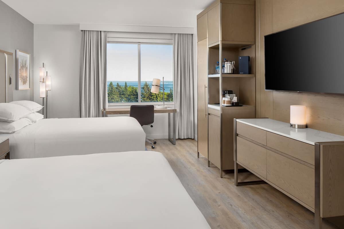 A hotel room with two beds, a desk, a chair, a TV, and a view of the ocean.