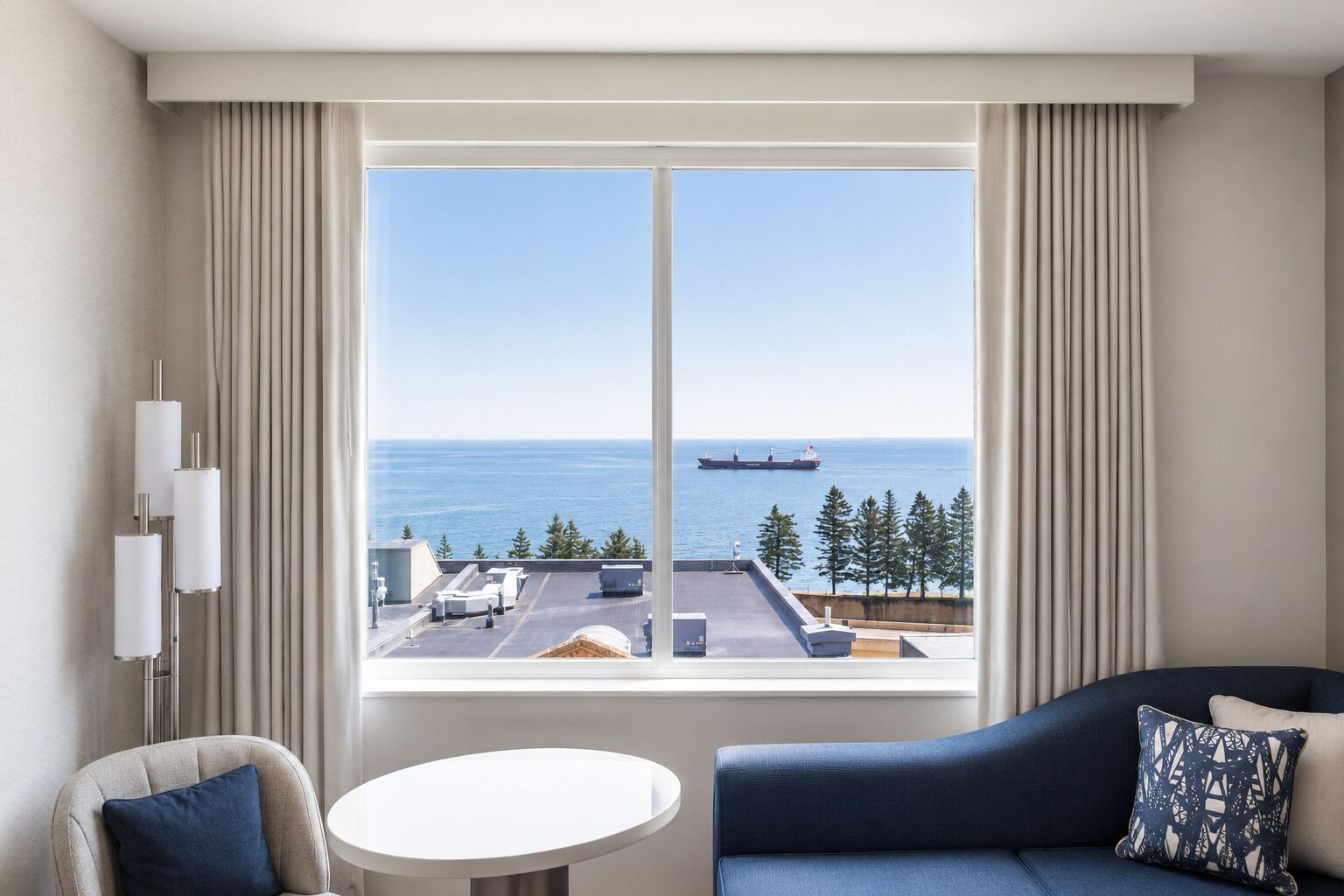 A hotel room with a large window offering a view of the sea and a ship, a round table, a sofa, and a chair.