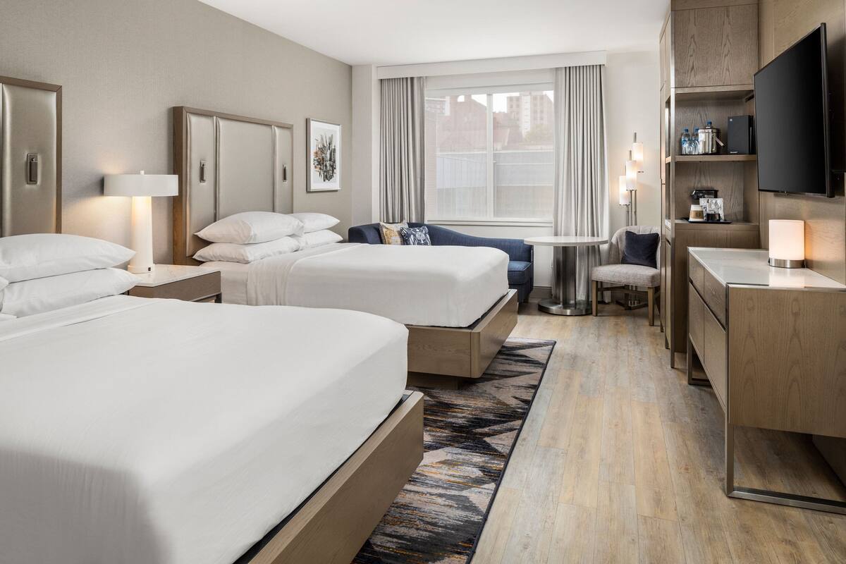 A hotel room with two beds, a desk, a chair, a TV, and a window with curtains.