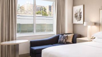 A hotel room with a bed, a sofa, a small table, a lamp, and a window with curtains.