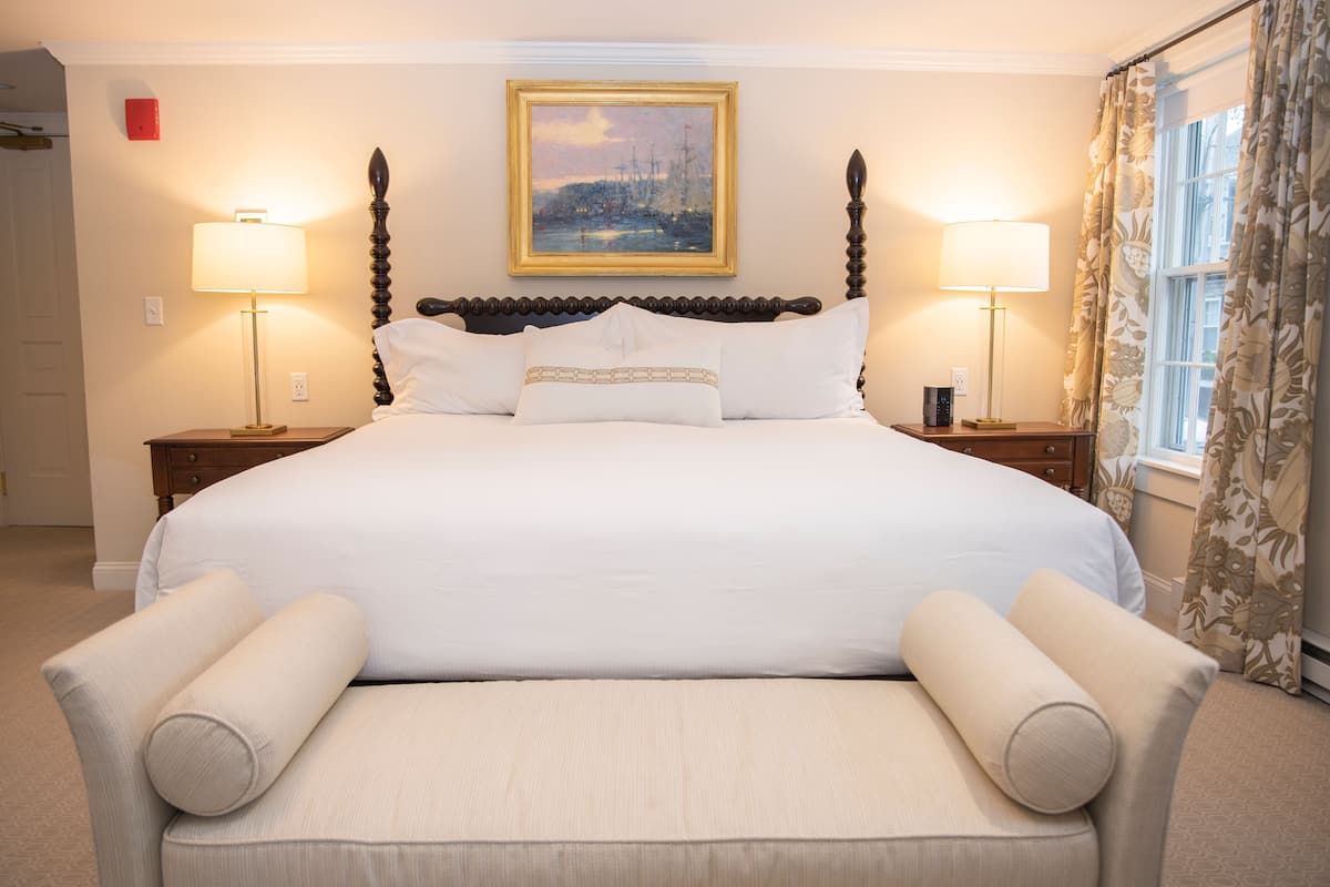 A large bed with a headboard, two bedside tables with lamps, a painting above the bed, and a window with curtains.