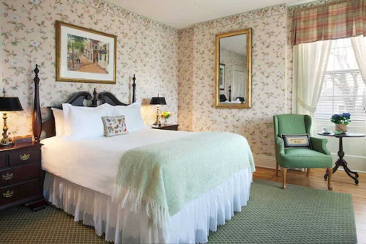 A neatly arranged bedroom with a large bed, a green armchair, a small table with a plant, and a mirror on the wall.