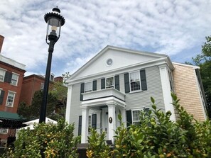 Une maison à deux étages, avec une façade blanche et des volets sombres, entourée de verdure et d’un lampadaire.