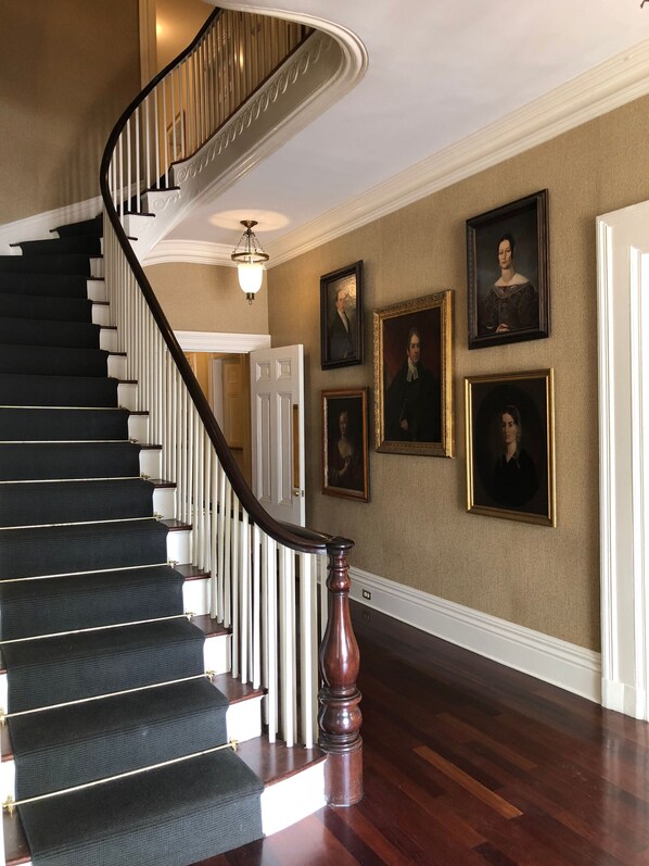 Una imponente escalera con acabado de madera oscura y balaustres blancos que conduce a una habitación con paredes color crema y retratos enmarcados.