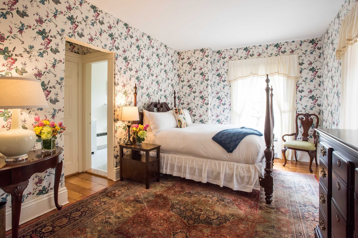 A bedroom with floral wallpaper, a four-poster bed, a nightstand with a lamp, a dresser, and a large rug.