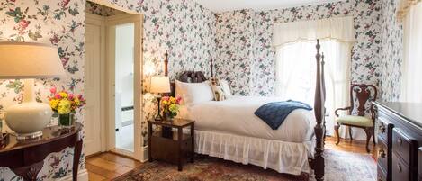 A bedroom with floral wallpaper, a four-poster bed, a nightstand with a lamp, a dresser, and a large rug.