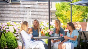 An outdoor patio with four people seated at a table, surrounded by striped umbrellas and floral arrangements. - Jared Coffin House (Nantucket)