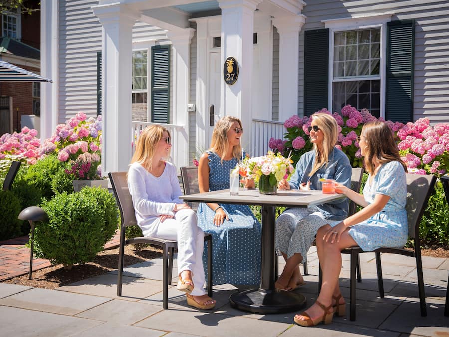 Une terrasse avec une table et des chaises, entourée de magnifiques parterres de fleurs et d’un porche blanc.