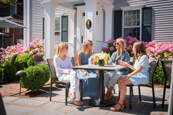 A patio with a table and chairs, surrounded by lush flower beds and a white porch. - Jared Coffin House (Nantucket)