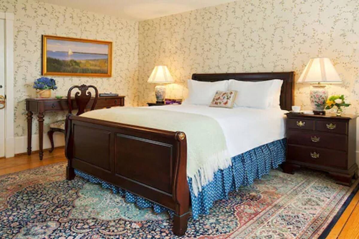 A bedroom with a large bed, two bedside tables with lamps, a nightstand, a chair, and a desk with a lamp and a vase of flowers.