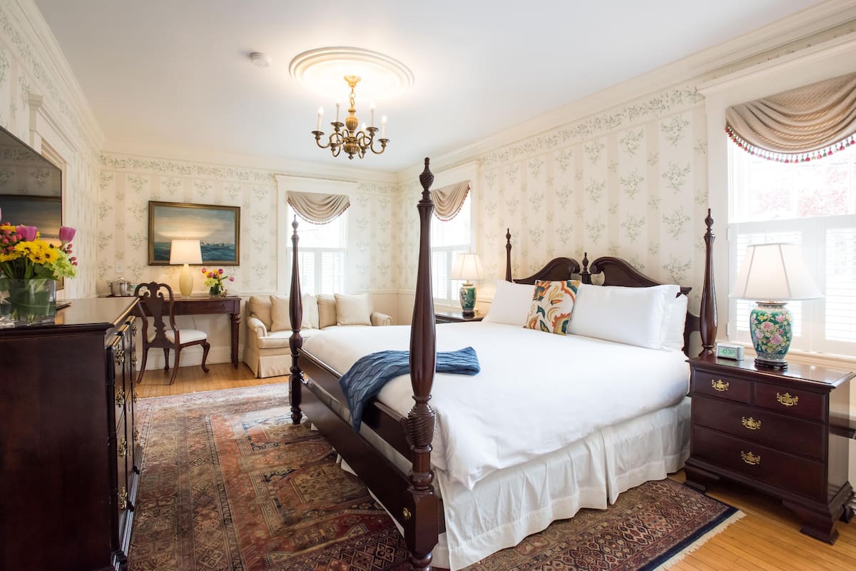 A four-poster bed with a white bedspread, a wooden nightstand with drawers, a desk with a chair, and a large window with curtains.