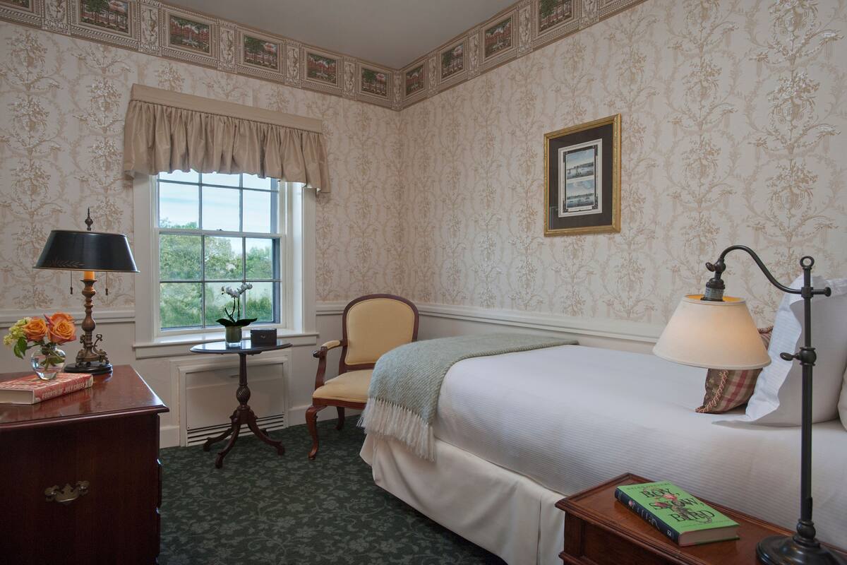 A bedroom with a bed, a chair, a desk with a lamp, a window, and a framed picture on the wall.