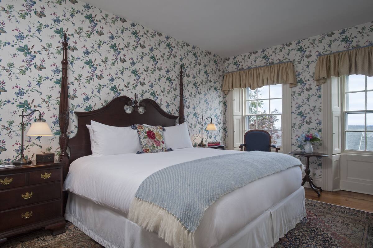 A bedroom with a large bed, floral wallpaper, a wooden nightstand, a chair, and a window with curtains.