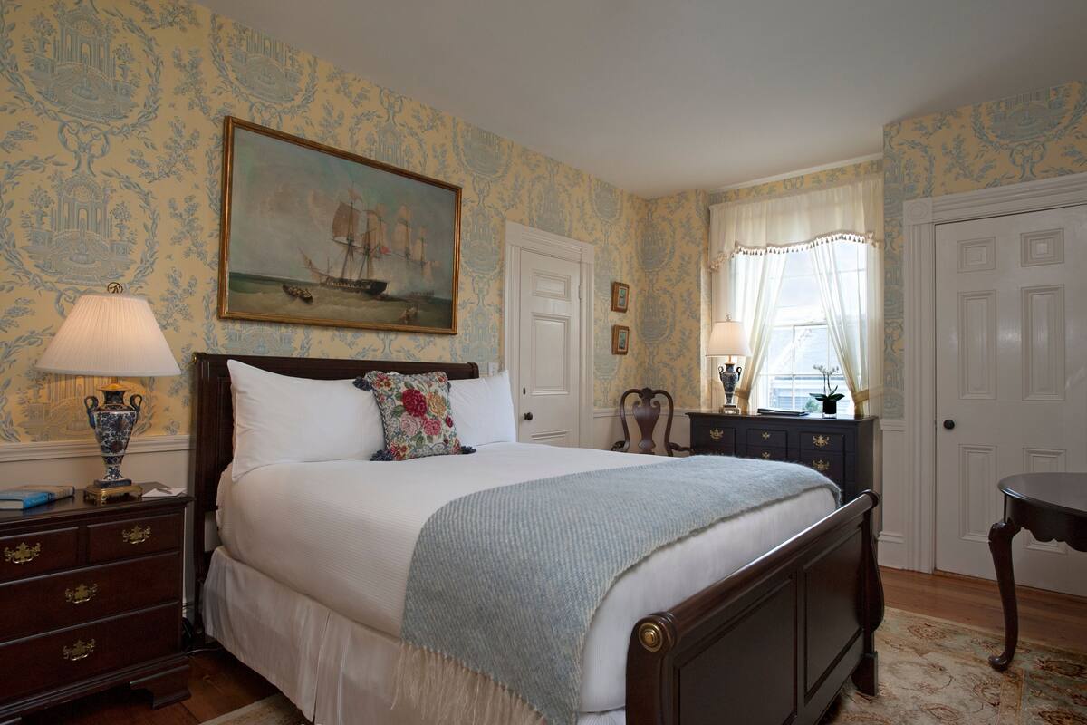 A neatly arranged bedroom with a large bed, a dresser, a nightstand, a painting, and a window with curtains.