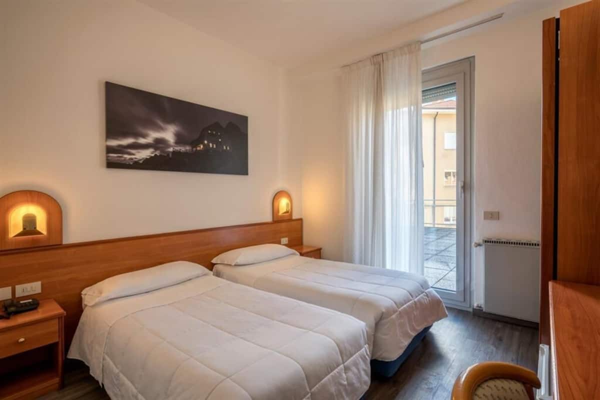 Une chambre d’hôtel avec deux lits, une table de chevet, une lampe, un tableau et une porte donnant sur un balcon.