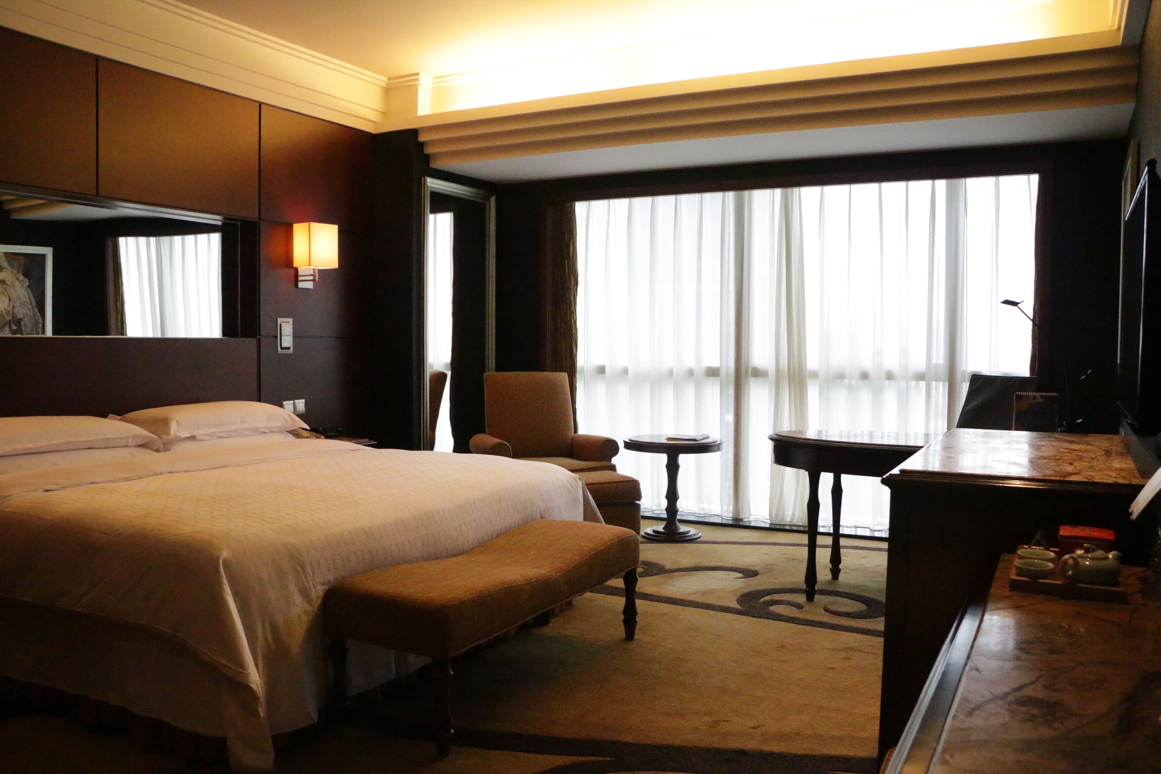 Ein Hotelzimmer mit einem großen Bett, einem Sessel, einem kleinen Tisch und einem Schreibtisch mit Fernseher.