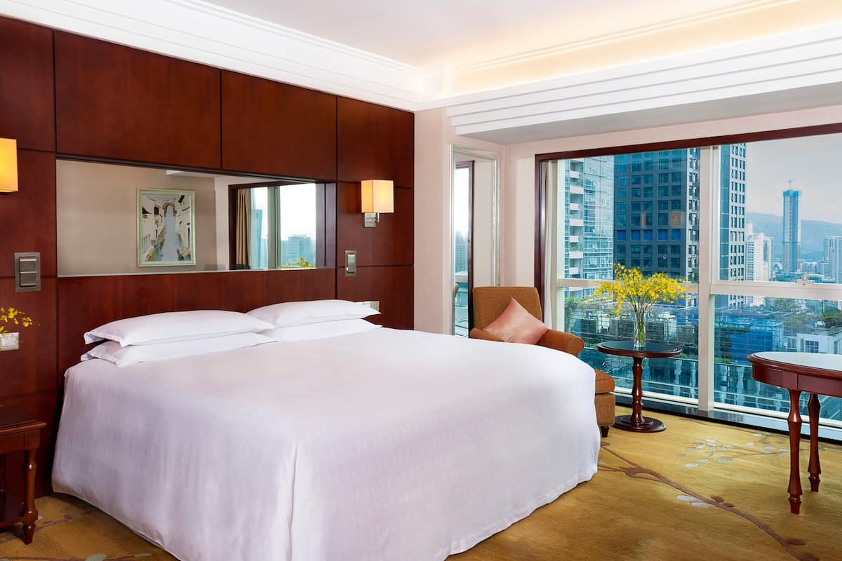 Ein Hotelzimmer mit einem großen Bett, einer Sitzecke mit Sessel und Tisch, einem großen Fenster mit Blick auf die Stadt und einem gerahmten Kunstwerk an der Wand.