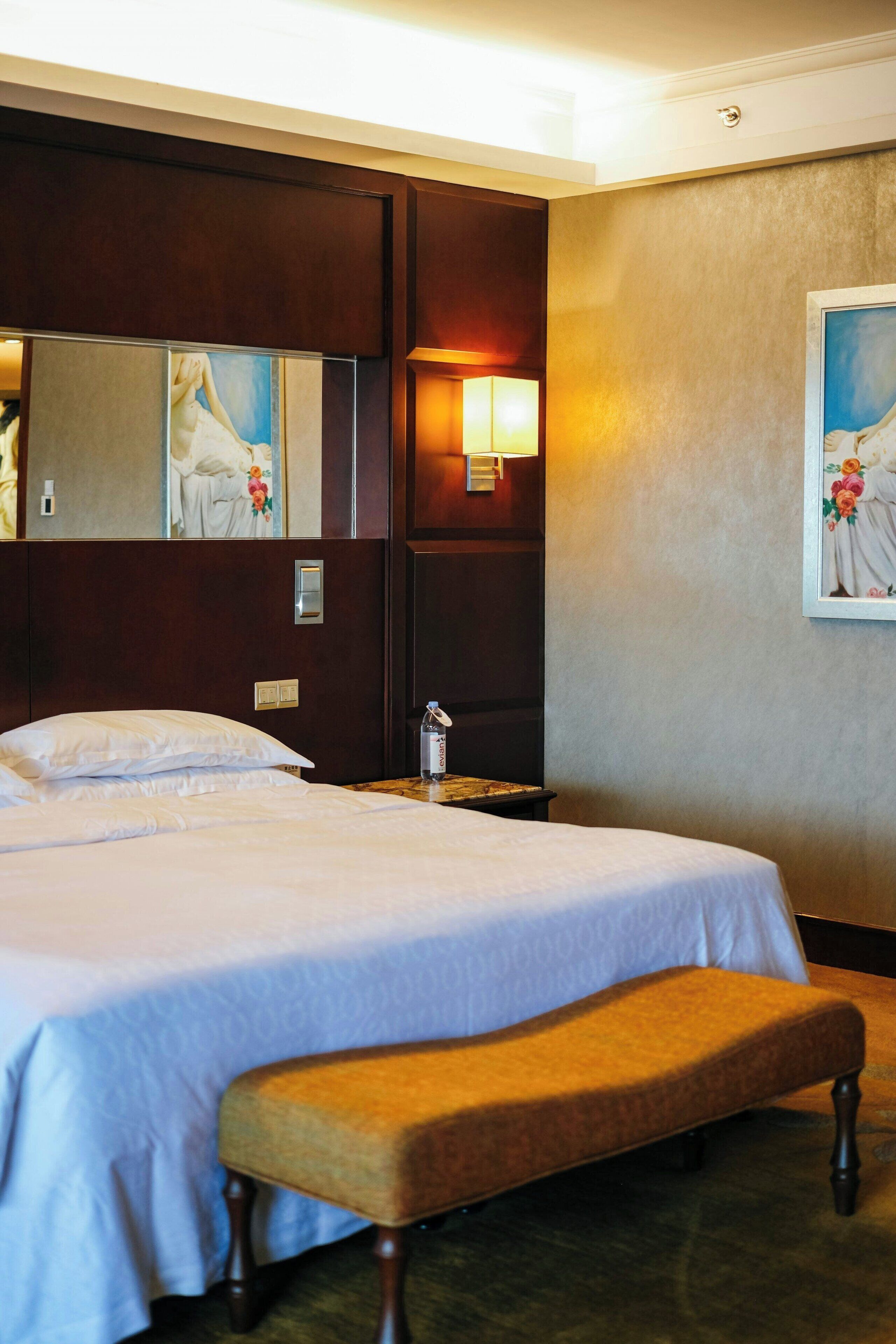A hotel room with a large bed, a bench, a nightstand, a lamp, and a painting on the wall.