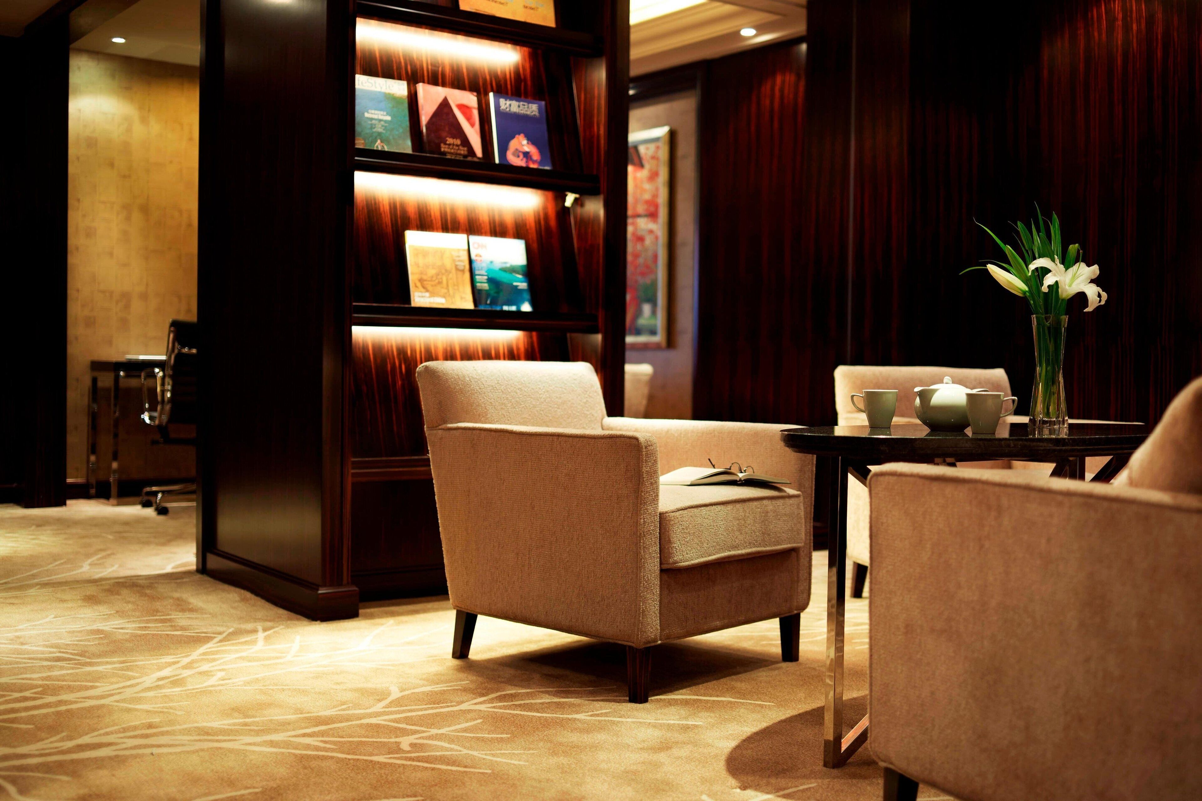 A hotel lobby with a beige armchair, a small table with a vase of flowers, and a bookshelf with framed pictures.