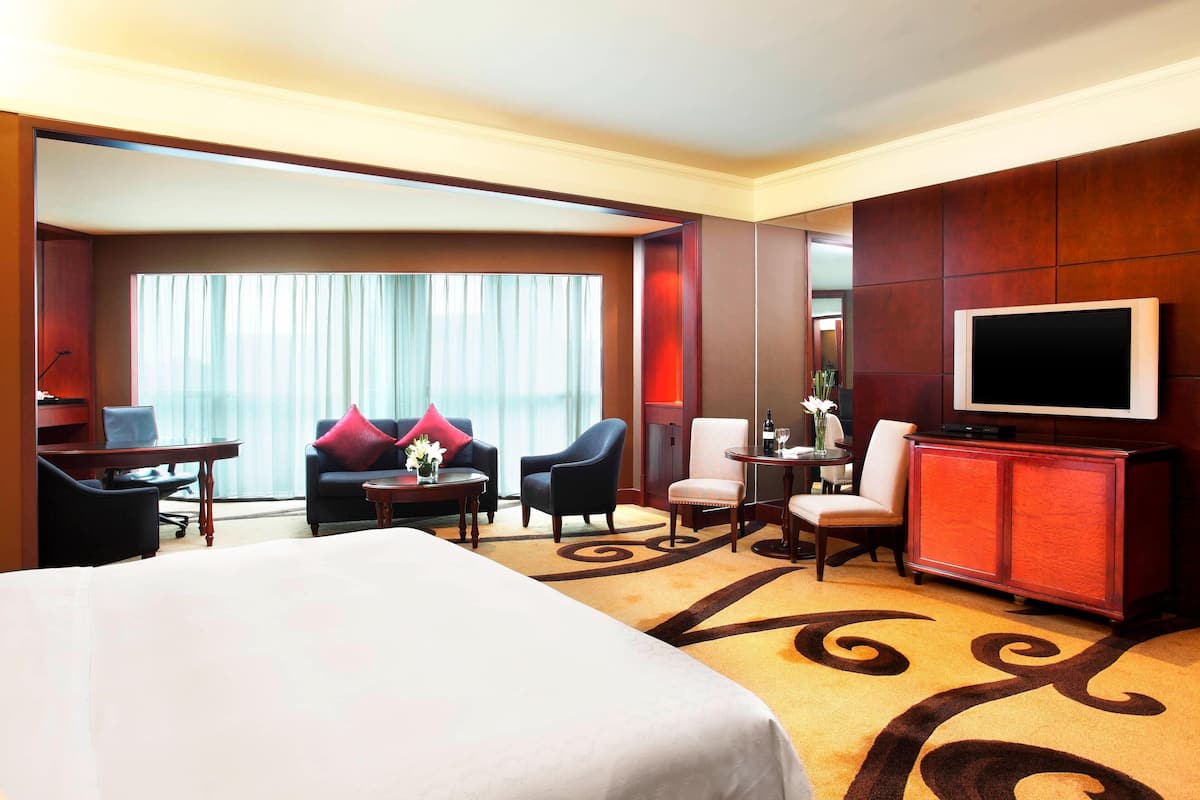 Ein Hotelzimmer mit einem großen Bett, einem Fernseher, einem Schreibtisch mit Lampe, zwei Sesseln, einem kleinen Tisch mit einer Vase und einem großen Fenster mit Vorhängen.