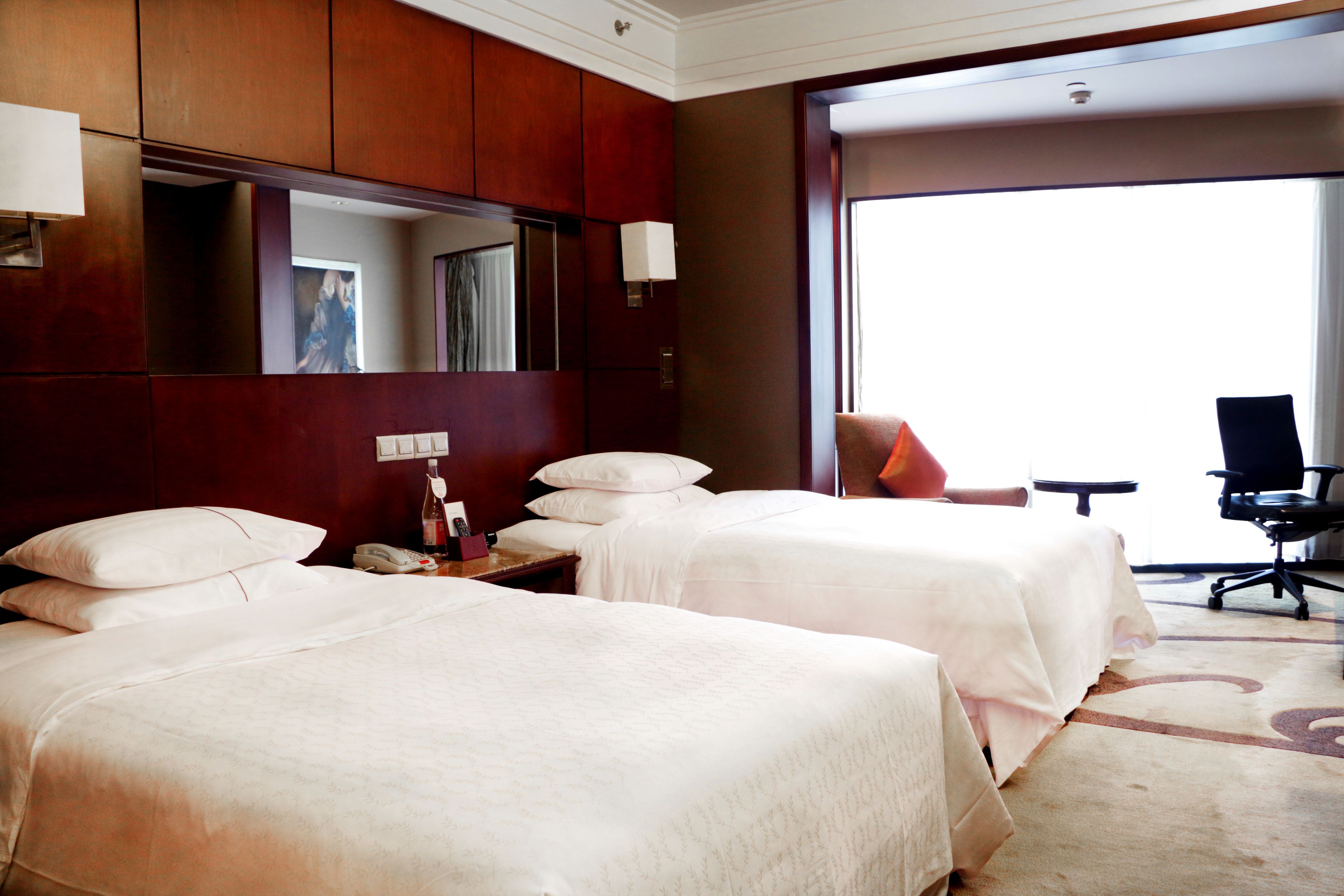 A hotel room with two beds, a desk with a lamp, a chair, and a large window.