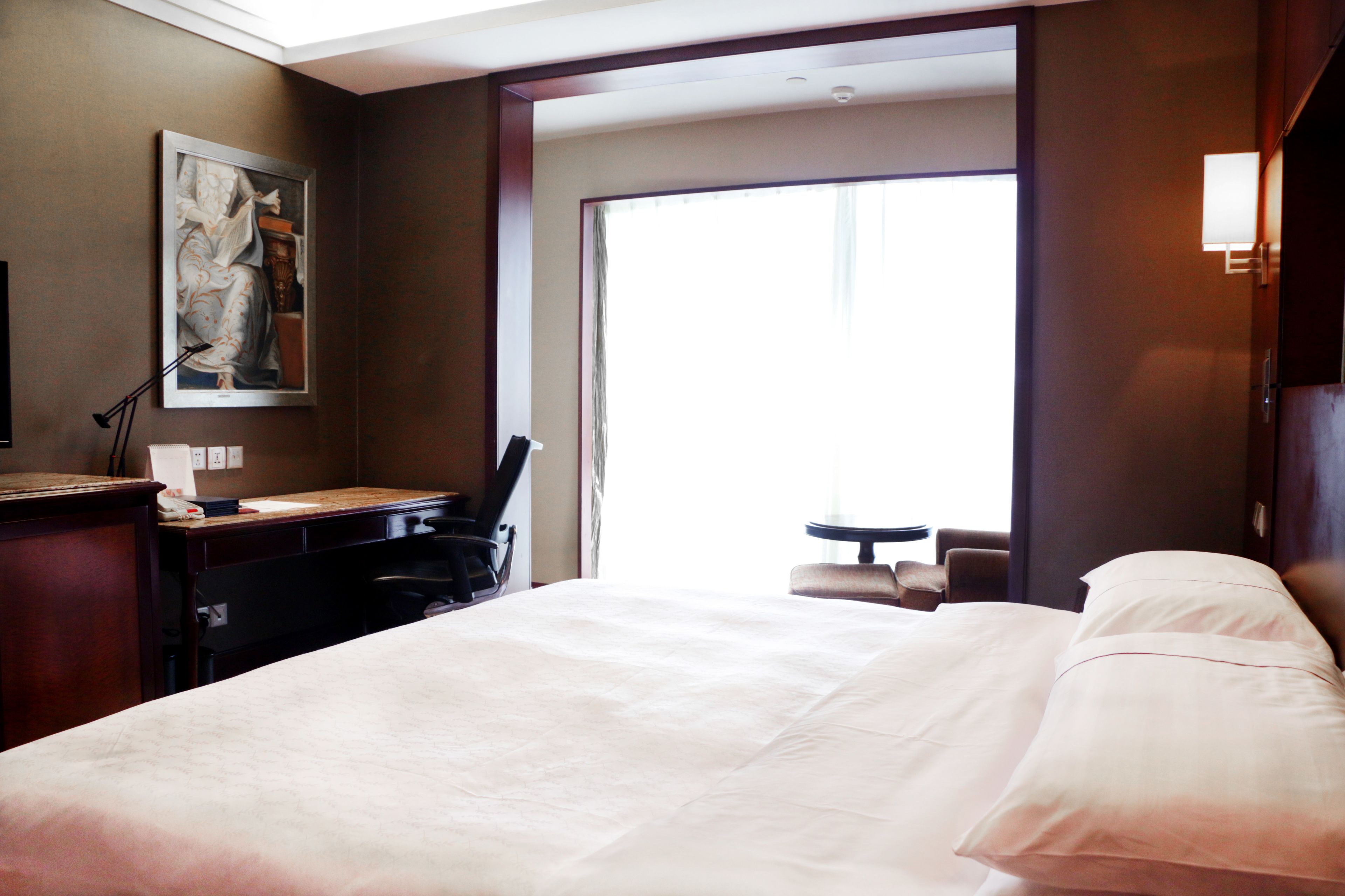 A hotel room with a large bed, a desk with a chair, a small table, and a painting on the wall.