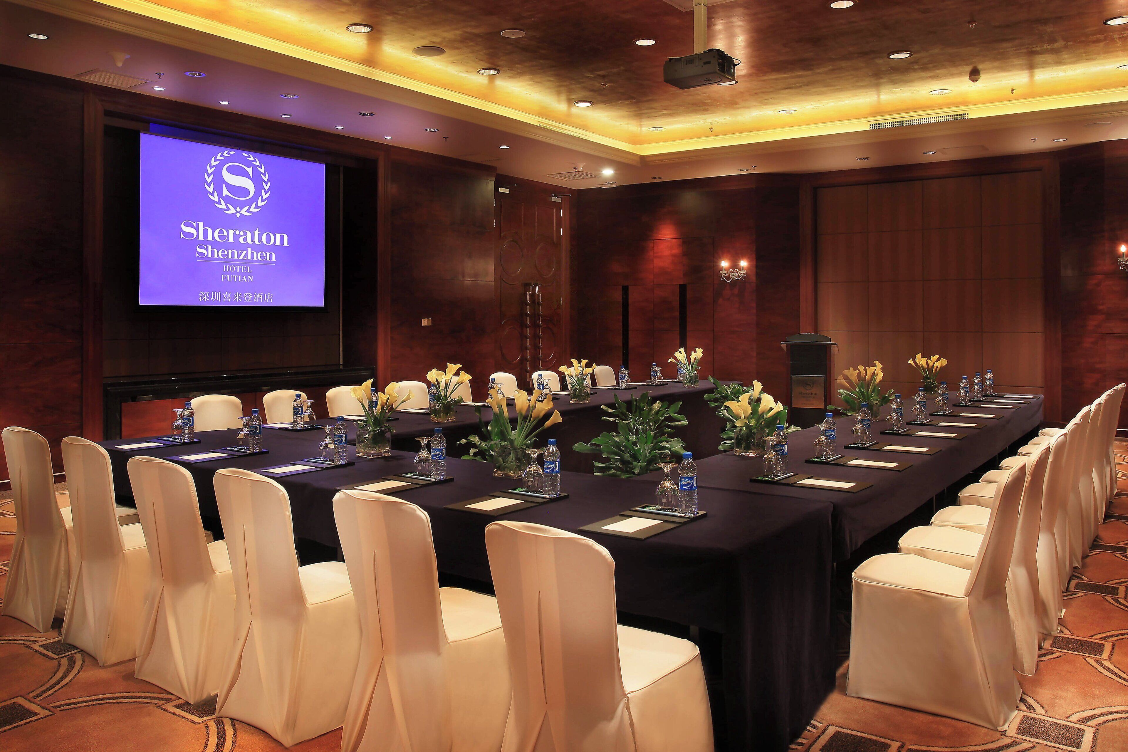 A conference room with a long table, chairs, and a screen displaying hotel information.