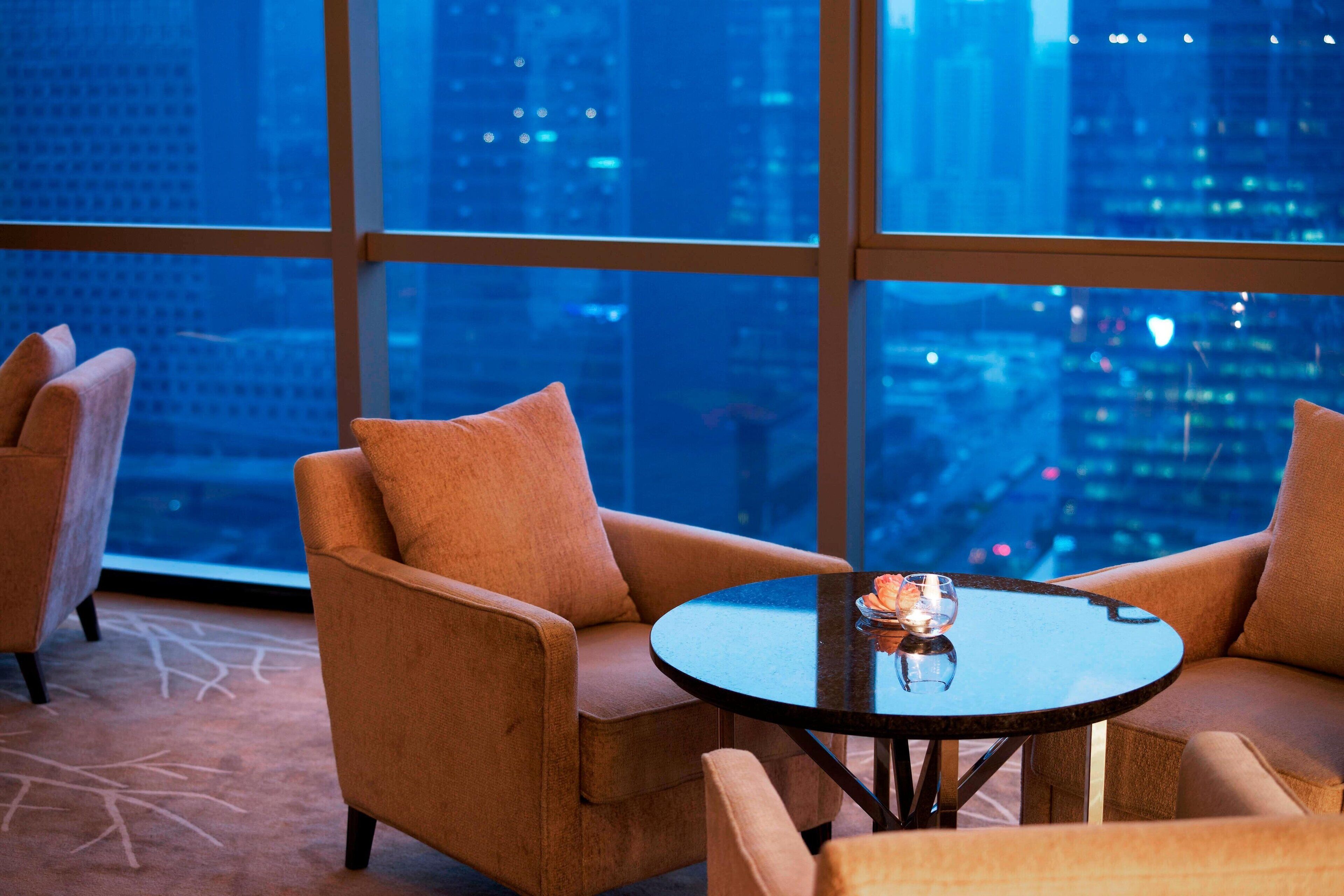 A modern hotel room with a large window offering a city view. There are two armchairs and a small round table with a glass.