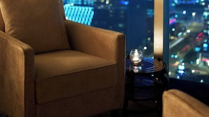 Een kamer met een groot raam met uitzicht op de stad 's nachts, een comfortabele fauteuil en een klein tafeltje met een decoratief object.