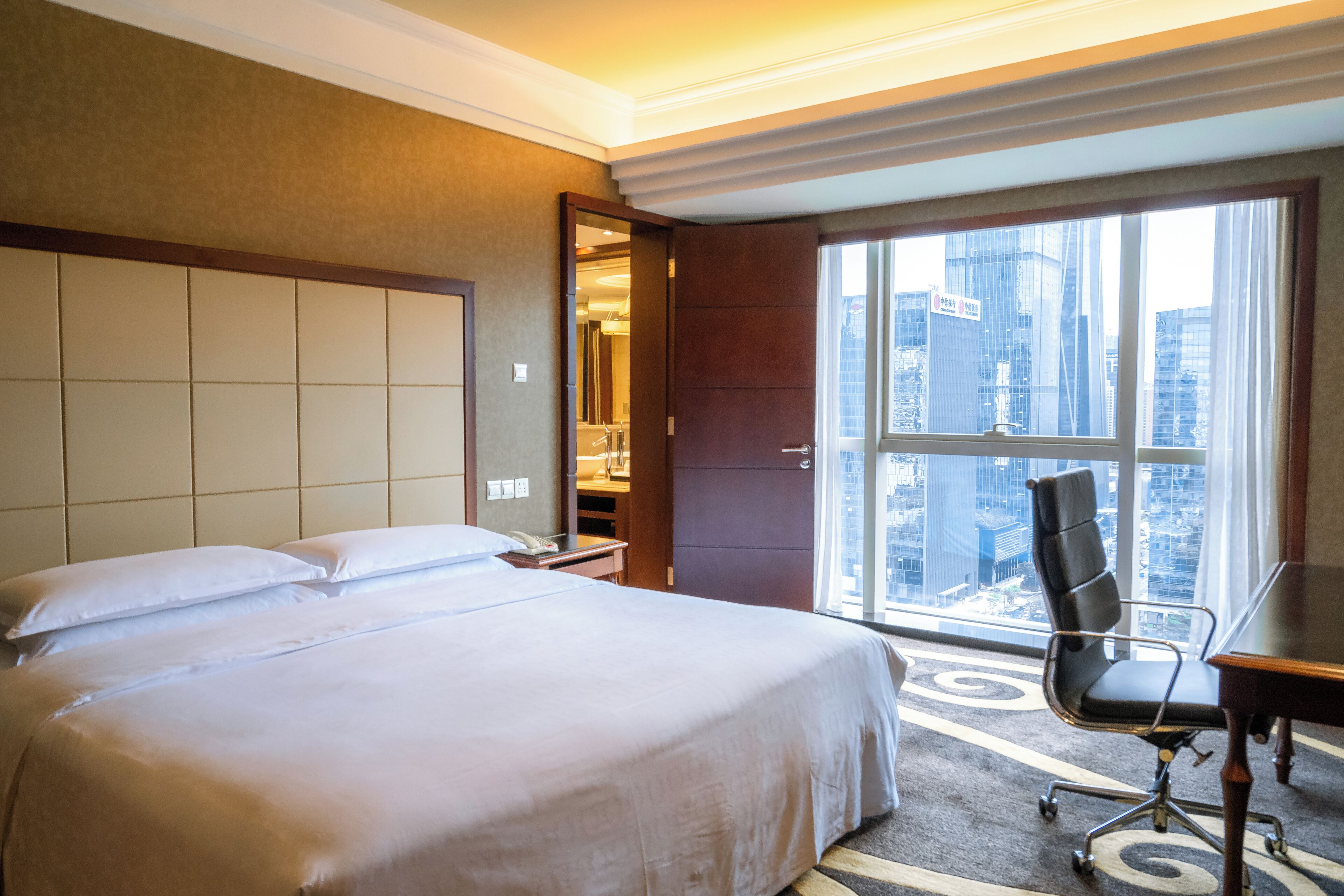 A hotel room with a large bed, a desk, and a view of a cityscape through a large window.