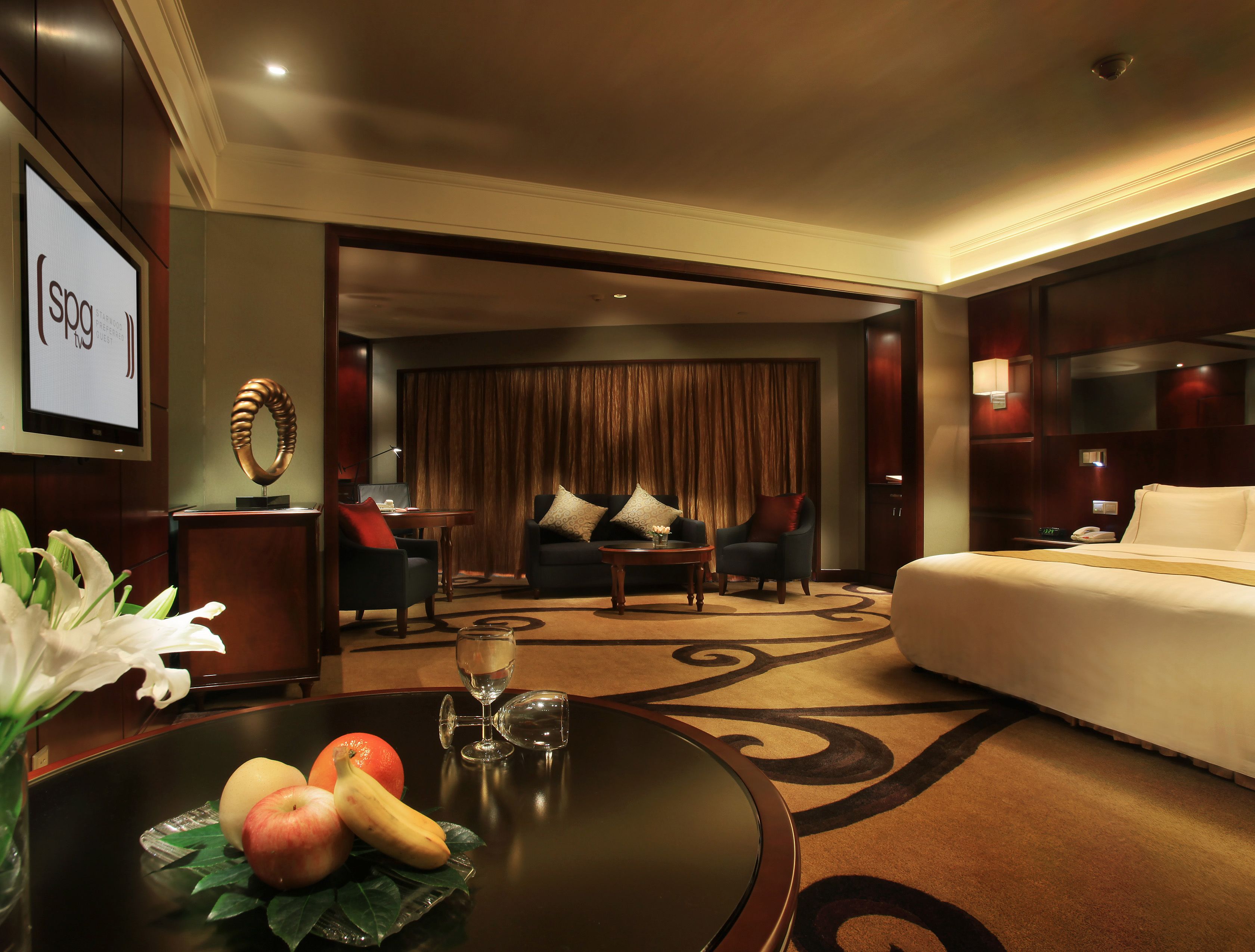 A hotel room with a bed, two armchairs, a sofa, a coffee table, and a TV screen displaying 'SPOT'.