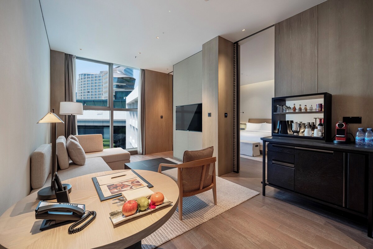 Una habitación de hotel moderna con sofá, una mesa de comedor con frutas, una cafetera y un amplio ventanal con vistas a la ciudad.