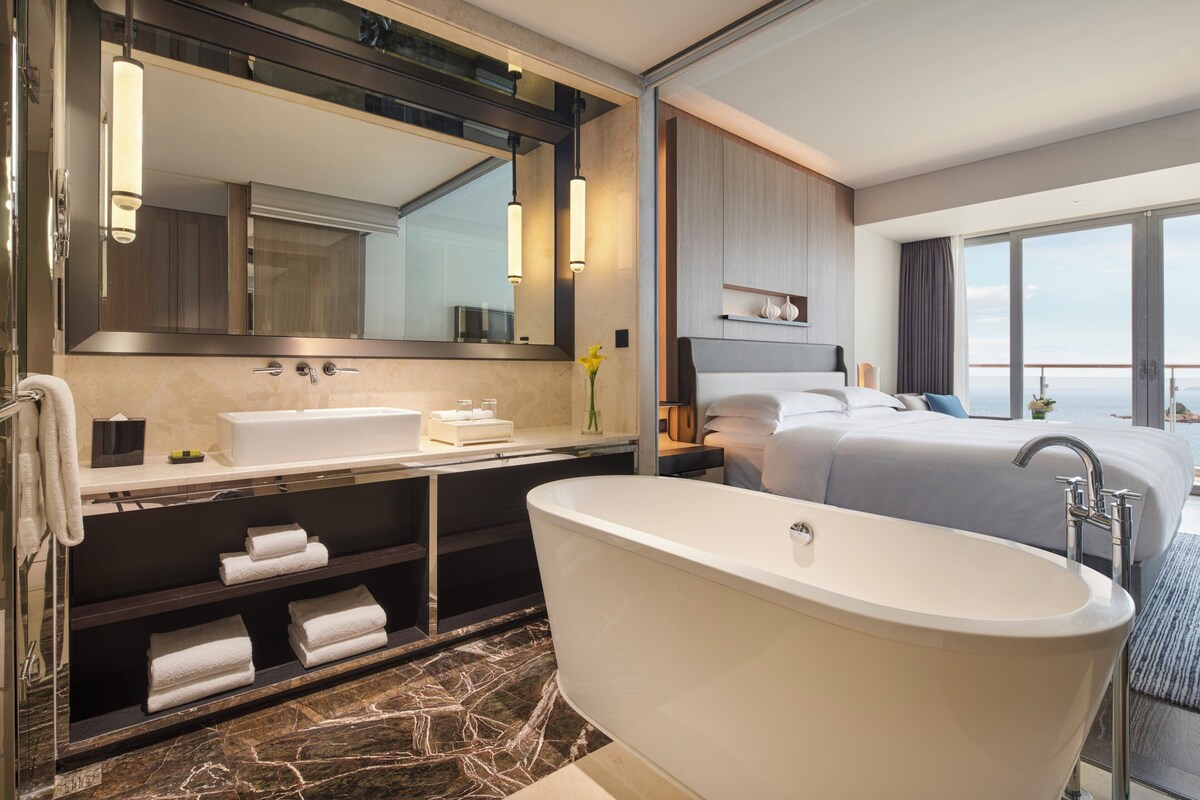 Un baño de hotel moderno con una bañera independiente, un espejo grande y un tocador con lavamanos y espacio para toallas.