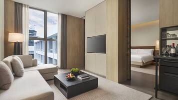 A modern hotel room with a sofa, a coffee table with fruits and books, a flat-screen TV, a bed, and a desk with a mirror and toiletries.