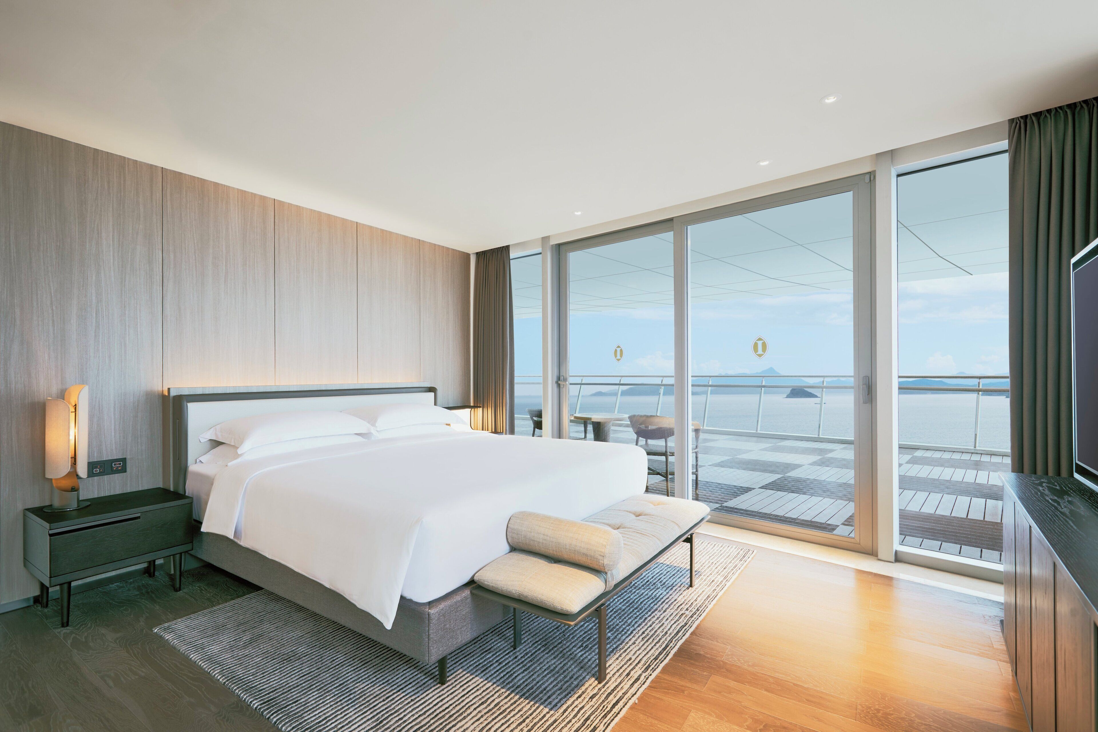 Un dormitorio moderno con una cama grande, una mesita de noche, un banco y vistas al mar a través de amplias ventanas.