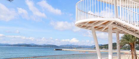 A seaside promenade with a white curved structure, a metal railing, and a clear view of the sea and distant mountains.