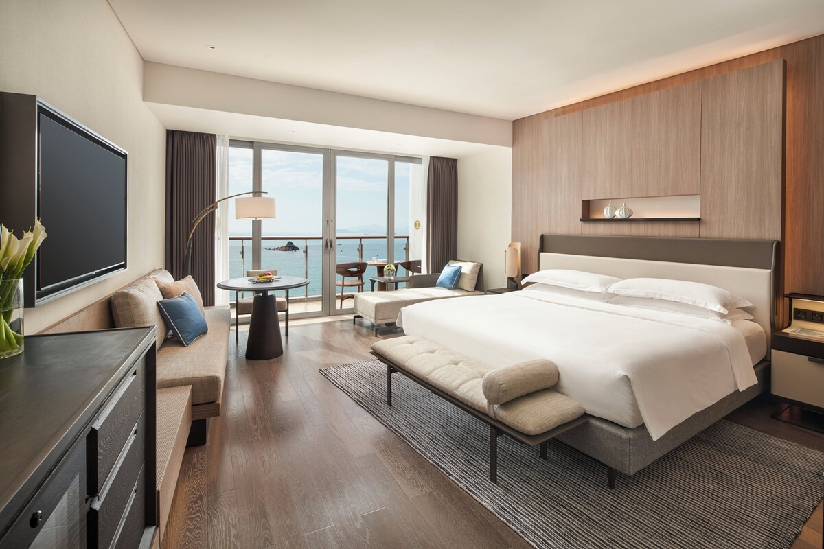 Una habitación de hotel moderna con una cama grande, un área de descanso con una mesa, un televisor y un balcón con vistas al mar.