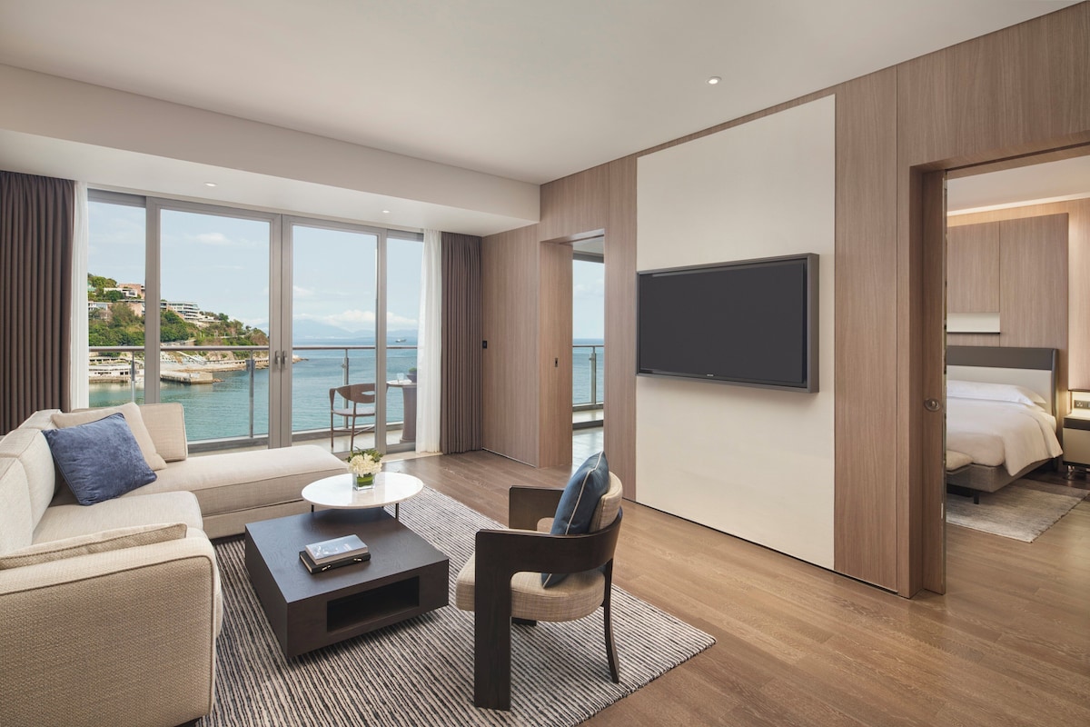 Habitación de hotel moderna con un amplio ventanal que ofrece vistas al mar y a las colinas, un televisor de pantalla plana montado en la pared, un sofá, una silla y una pequeña mesa de centro.