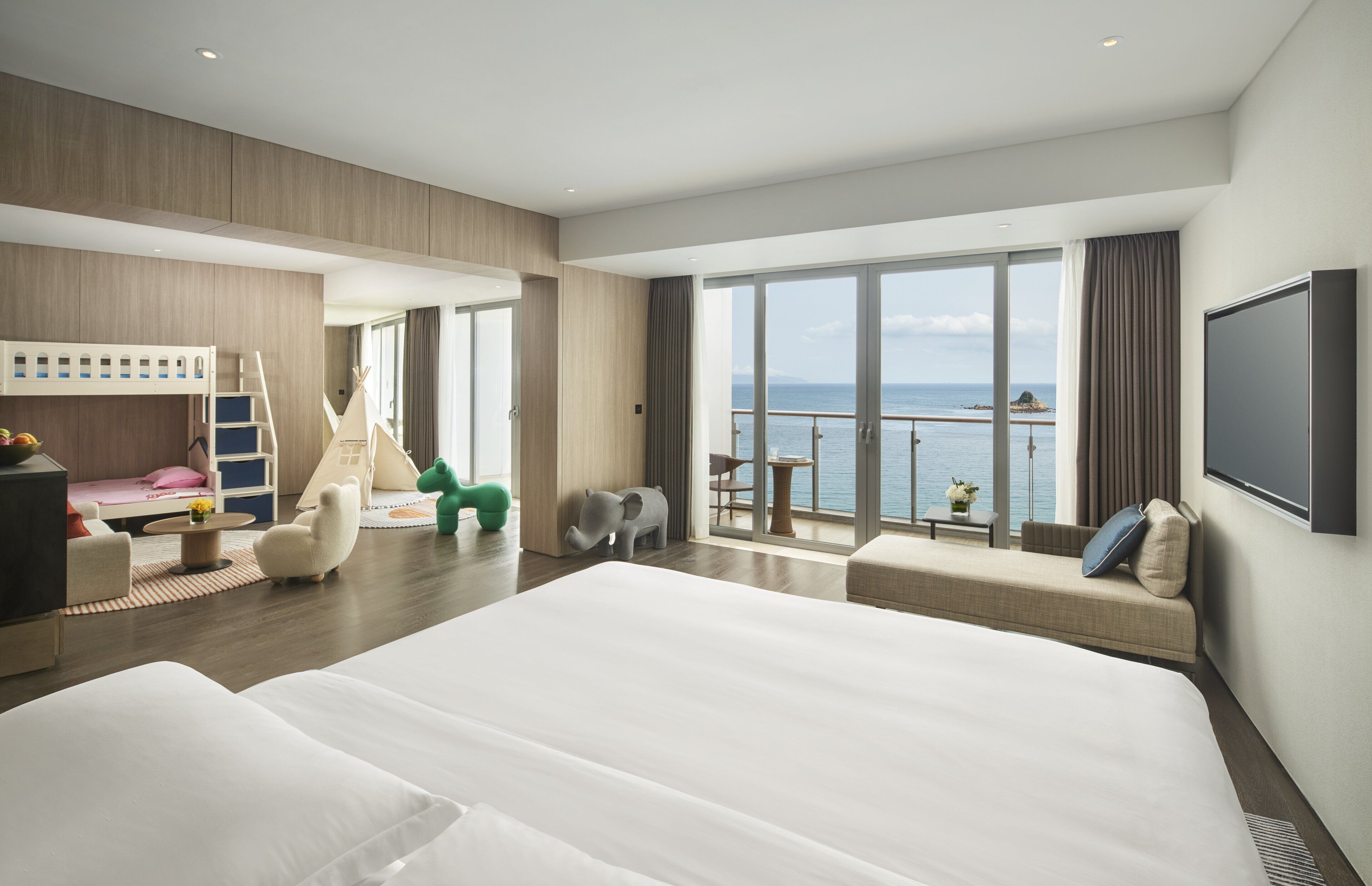 A modern hotel room with a large bed, a seating area with a view of the sea, a small table, and a TV.