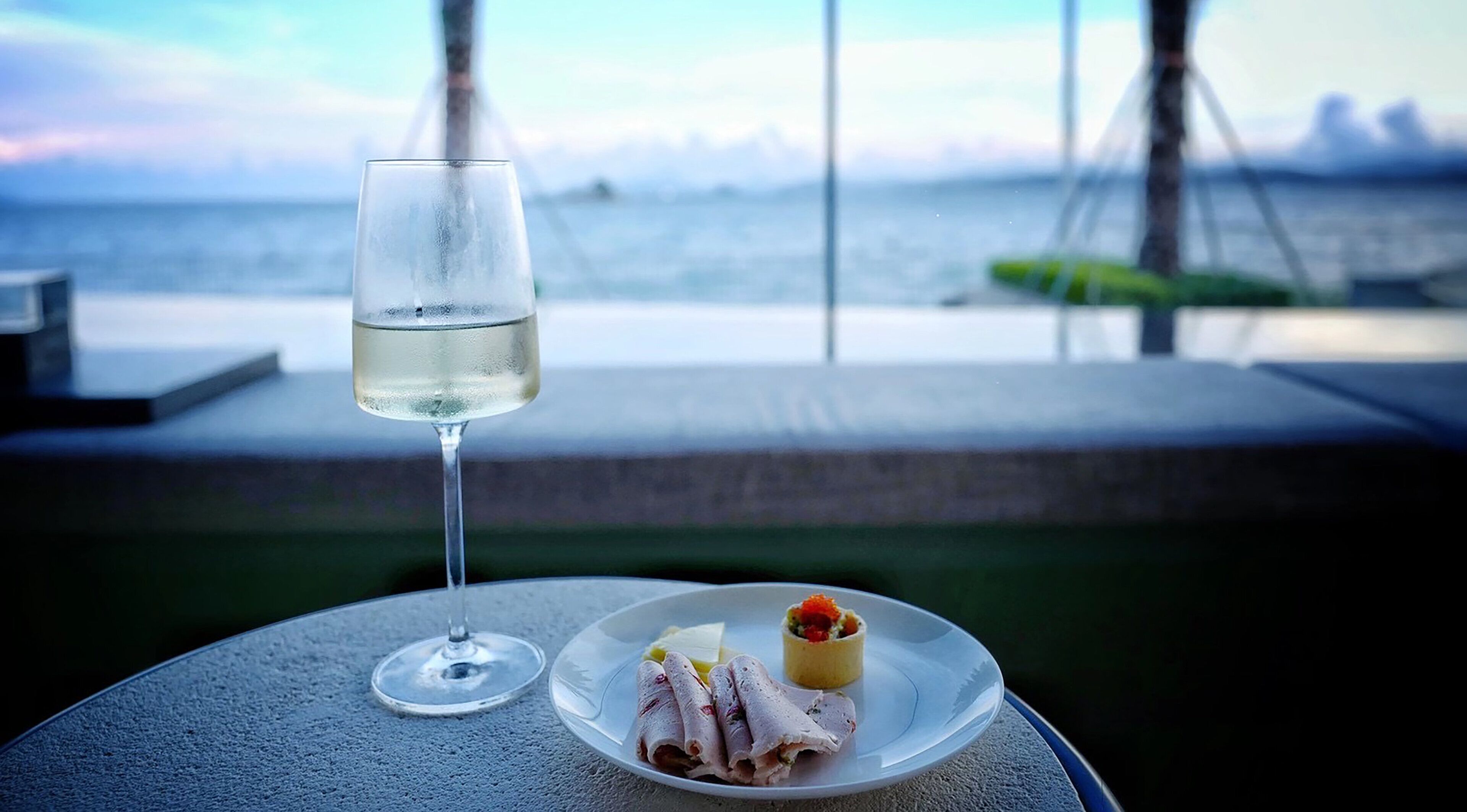 A glass of white wine on a table with a plate of food and a scenic view of the sea and mountains in the background.