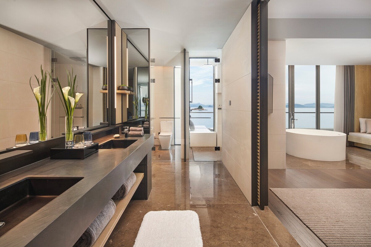 Un baño moderno con un espejo grande, un lavamanos y un inodoro. Se ve el mar a través de la ventana del baño.