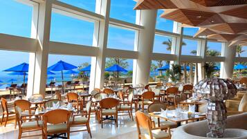 A spacious outdoor dining area with numerous tables and chairs, large windows, and blue umbrellas.