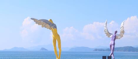 Una playa con dos grandes estatuas de ángeles, una amarilla y otra púrpura, sobre la arena, con vista despejada al mar y montañas distantes.