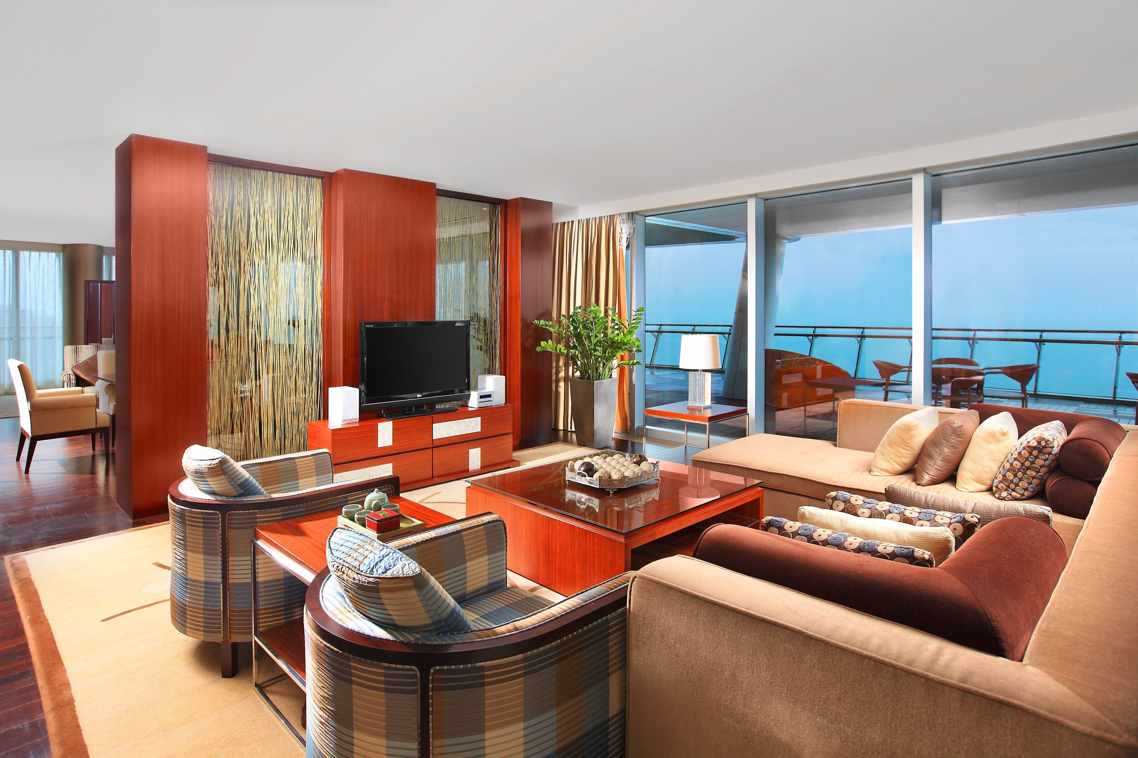 A modern hotel room with a large flat-screen TV, a sofa set, a dining table, and a balcony with a view of the sea.