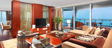 Quarto de hotel moderno com televisão de tela plana grande, um sofá, uma mesa de jantar e uma varanda com vista para o mar.