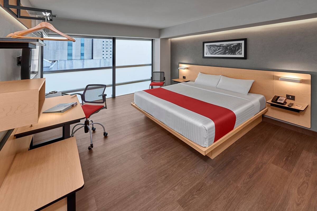 A modern hotel room with a large bed, a desk with a chair, a desk lamp, a telephone, and a window with a view of buildings.