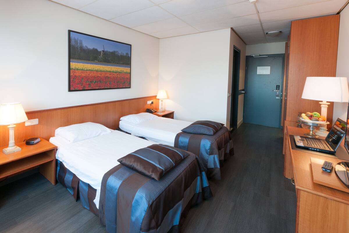 Ein Hotelzimmer mit zwei Betten, einem Schreibtisch mit einem Laptop, einer Lampe und einem Bild an der Wand.